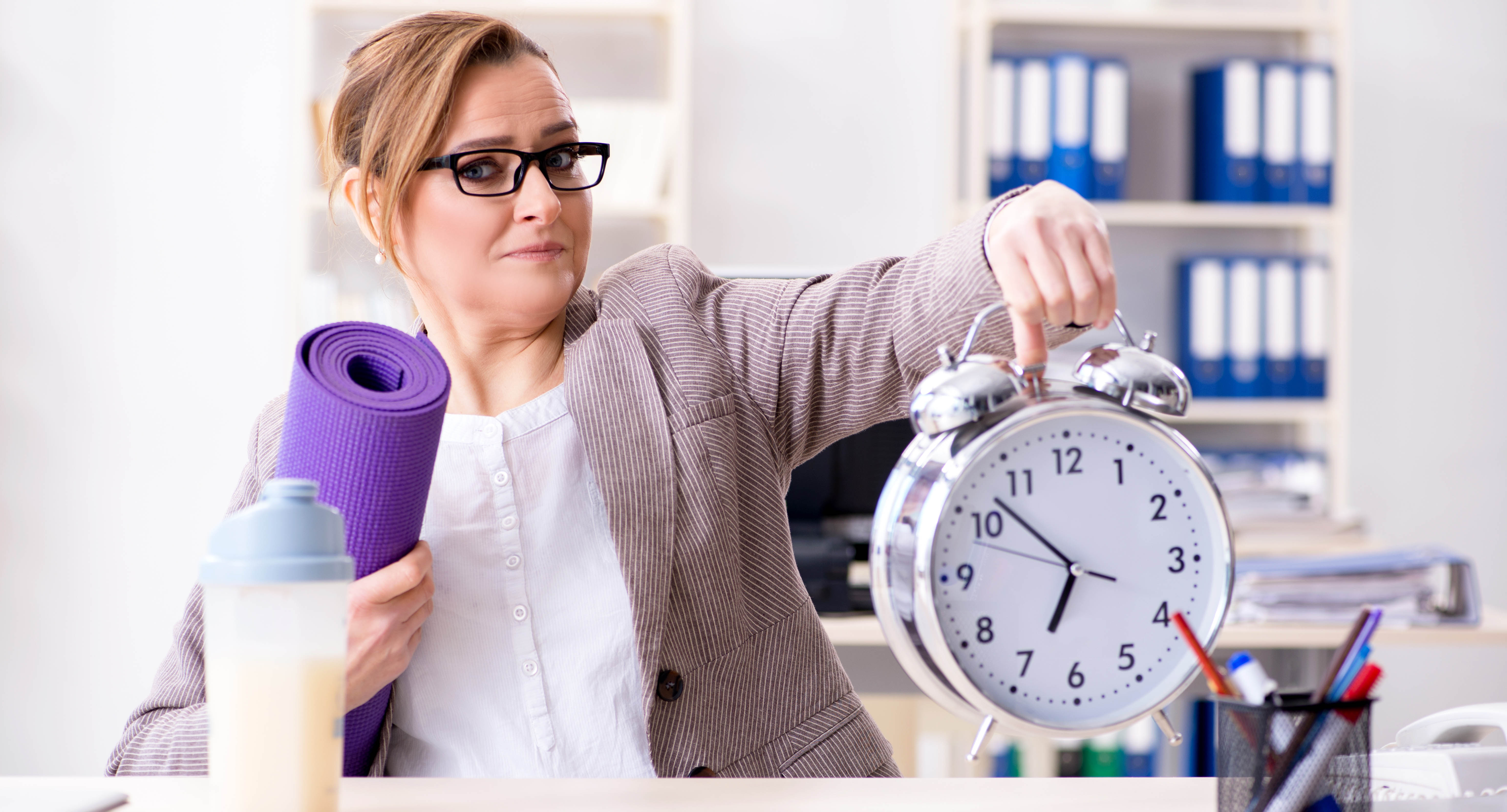 Professional woman sitting at her desk holding a large alarm clock in one hand and her purple yoga mat in the other, preparing to go to the gym for a workout